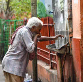 An old person drinks water from a small outdoor trough