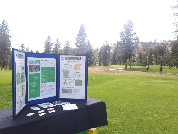 A table on a golf course green features an information display for the Thompson Okanagan Tourism Association's Golf Tournament 'drive' for sustainability.